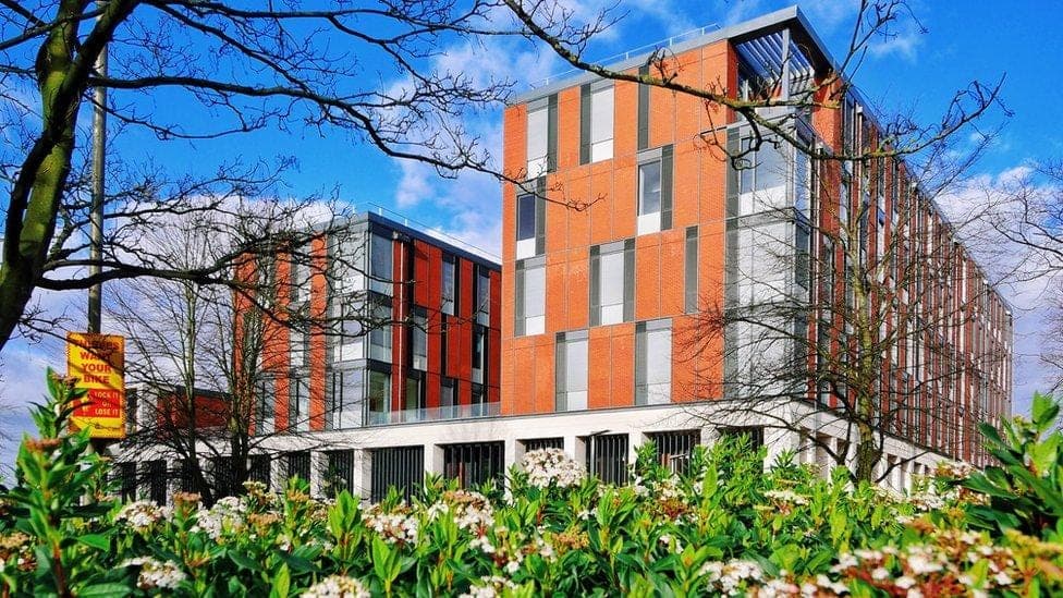 The George Davies Centre, a modern building with orange and red panels and large windows, seen behind trees and flowering shrubs.